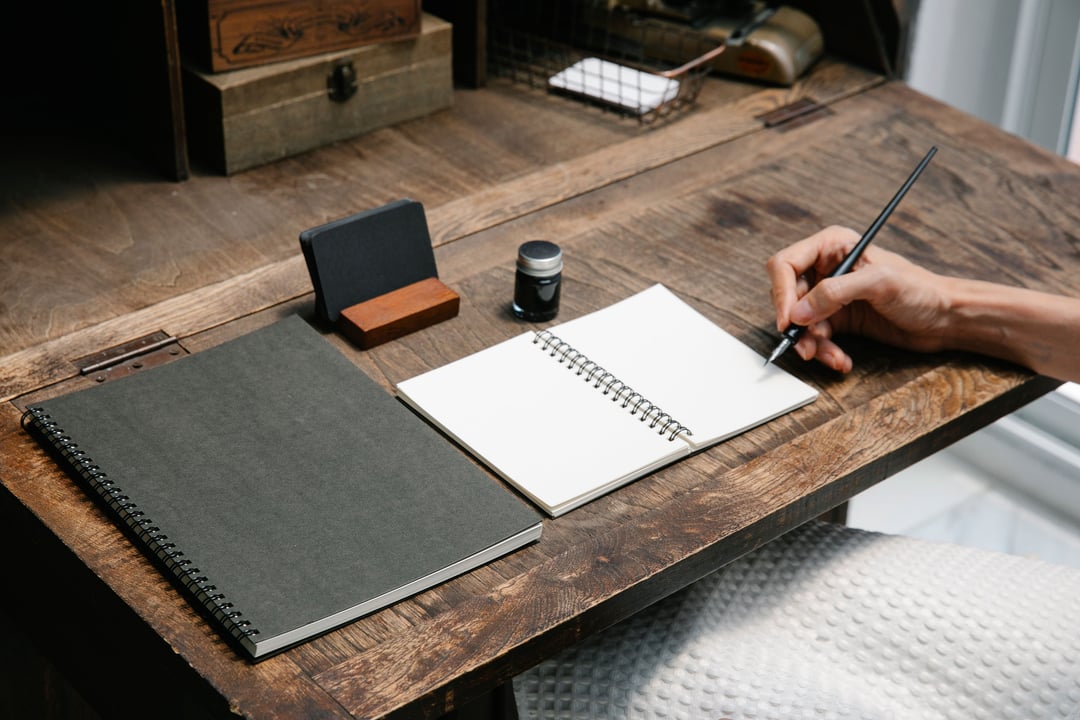 A hand writing in an open notebook on a wooden desk.